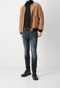Veste bomber en daim marron, col roulé noir, jean denim foncé et chaussures à lacets noires brillantes. Caractéristiques : poignets côtelés et poche zippée.