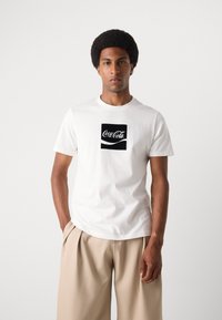 Witte katoenen t-shirt met een zwart Coca-Cola logo. Korte mouwen en een ronde halslijn, gedragen met beige geplooide broek.