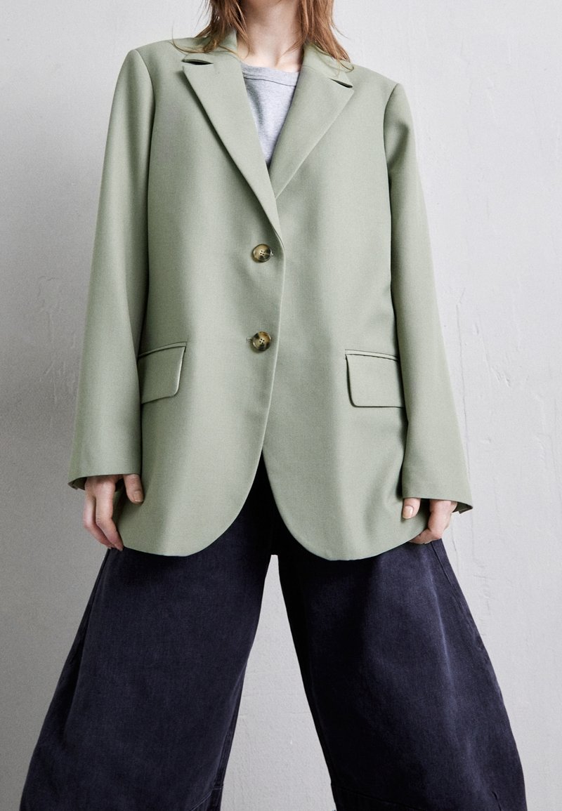 Blazer verde chiaro con due bottoni, rever classici e tasche frontali. Presenta una vestibilità strutturata e un tessuto liscio e testurizzato.