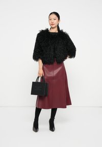 Veste en fausse fourrure noire coupée, jupe en cuir bordeaux, sac à main noir avec des accents cloutés, collants noirs et chaussures à bout pointu, pose debout.