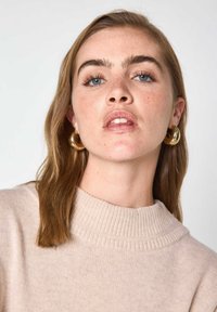 Maglione beige con colletto alto a coste. Il modello ha i capelli castano chiaro e indossa grandi orecchini a cerchio dorati. Pelle chiara con lentiggini.
