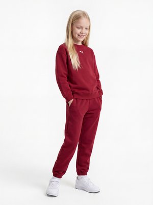 Młoda dziewczyna z długimi blond włosami, ubrana w czerwony dres Puma i białe trampki, stoi z rękami w kieszeniach i się uśmiecha.