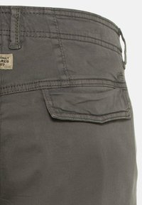 Graue Stoffhose mit strukturiertem Oberflächenmaterial, mit einer Gesäßtasche und dezenten Nähdetails. Etikett über der Gesäßtasche angebracht.