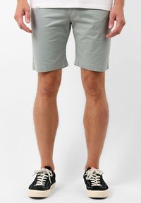 Shorts en coton vert clair à coupe droite, avec ourlet retroussé et poches latérales, accompagnés de baskets noires avec des lacets et des semelles blanches.