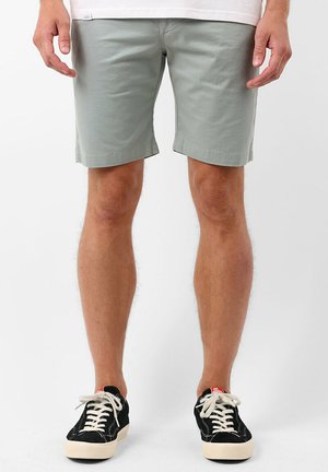 Shorts en coton vert clair à coupe droite, avec ourlet retroussé et poches latérales, accompagnés de baskets noires avec des lacets et des semelles blanches.