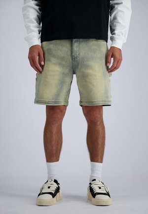 Mann trägt ausgewaschene Jeansshorts, weiße Sportsocken und schwarz-weiße Turnschuhe vor einfachem Hintergrund.