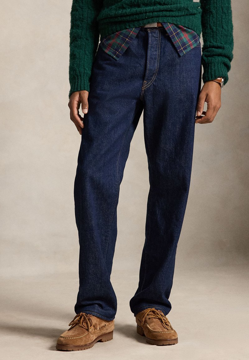 Persona con vaqueros de mezclilla azul oscuro, zapatos marrones de ante con cordones, suéter verde y camisa de cuadros debajo.