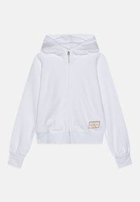 Sudadera blanca con cremallera, capucha, puños acanalados, un diseño ajustado y un pequeño parche de logo en un tono neutro en la parte inferior izquierda.