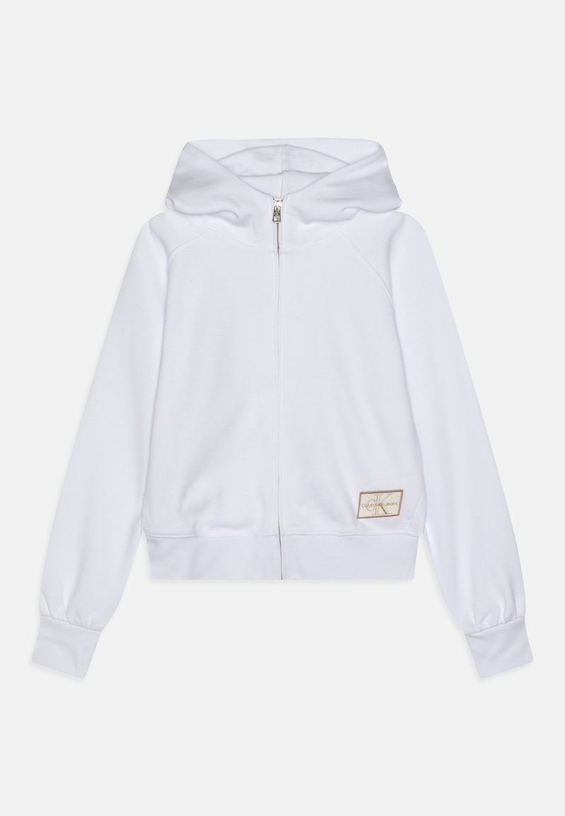 Sudadera blanca con cremallera, capucha, puños acanalados, un diseño ajustado y un pequeño parche de logo en un tono neutro en la parte inferior izquierda.