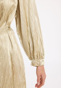 Robe longue à manches bouffantes en satin beige avec un motif texturé subtil. Elle possède une ceinture assortie et des poignets froncés. Fini lisse.
