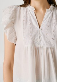 Λευκό μπλουζάκι με φουσκωτά μανίκια και κεντημένο με λουλουδάτα σχέδια στο άνω μέρος του στήθους, με κοντή σειρά κουμπιών και υφασμάτινη συγκέντρωση από κάτω.