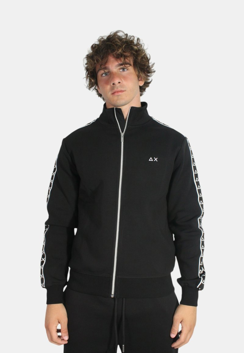 Felpa nera con zipper e colletto alto, con accenti bianchi a fantasia lungo le maniche, texture morbida e polsini aderenti. Logo minimalista sul petto.