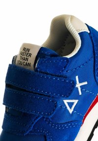 Scarpa sportiva blu con cinturini in Velcro e dettagli bianchi. Presenta un tessuto testurizzato e un piccolo etichetta con scritto "CORRI PIÙ VELOCE DI QUANTO PUOI."