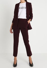 Burgundský blazer přes bílé tričko s nápisem "PARADIS", doplněný o sladěné slim kalhoty a černé vysoké podpatky. Hladká tkanina, upravený střih.