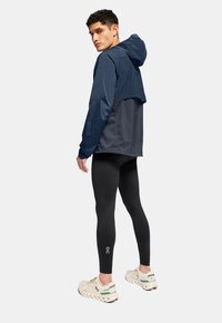 Water-resistant jacket in navy and gray, featuring a hood, elastic cuffs, and a lightweight design, paired with black leggings and sneakers.