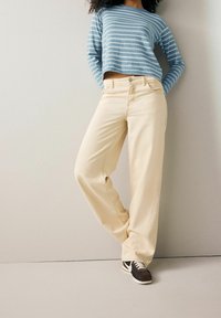 Licht beige wijde broek met voorzakken, gecombineerd met een cropped blauw-wit gestreept shirt met lange mouwen en bruine sneakers.