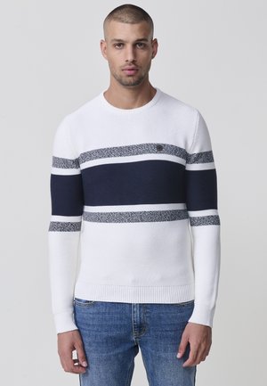 Pull en maille blanche avec des rayures bleu marine et texturées, col rond, et poignets et ourlet côtelés. Associé à un jean en denim bleu.