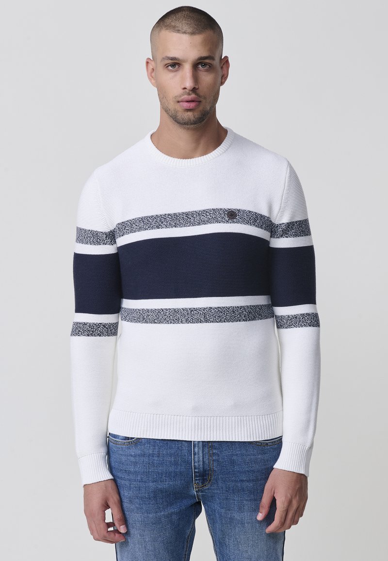 Weißer gestrickter Pullover mit marineblauen und strukturierten Streifen, Rundhalsausschnitt sowie gerippten Bündchen und Saum. Kombiniert mit blauen Jeans.
