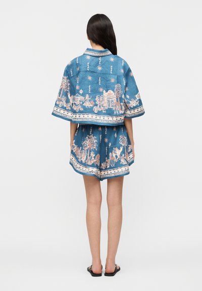 Γυναίκα που φοράει μπλε δίκομμα σύνολο με λευκά και ροζ περίπλοκα μοτίβα χωριού και φύσης, στέκεται με την πλάτη προς την κάμερα.