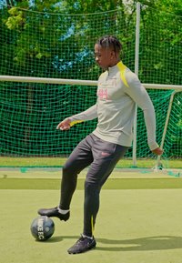Camiseta atlética gris con detalles amarillos, pantalones atléticos negros y botas de fútbol negras. Jugador haciendo malabares con un balón de fútbol negro en un campo sintético.