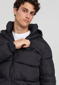 Giubbotto imbottito nero con tasca sul petto con zip, polsini elastici e colletto alto, caratterizzato da una texture lucida e un design trapuntato.