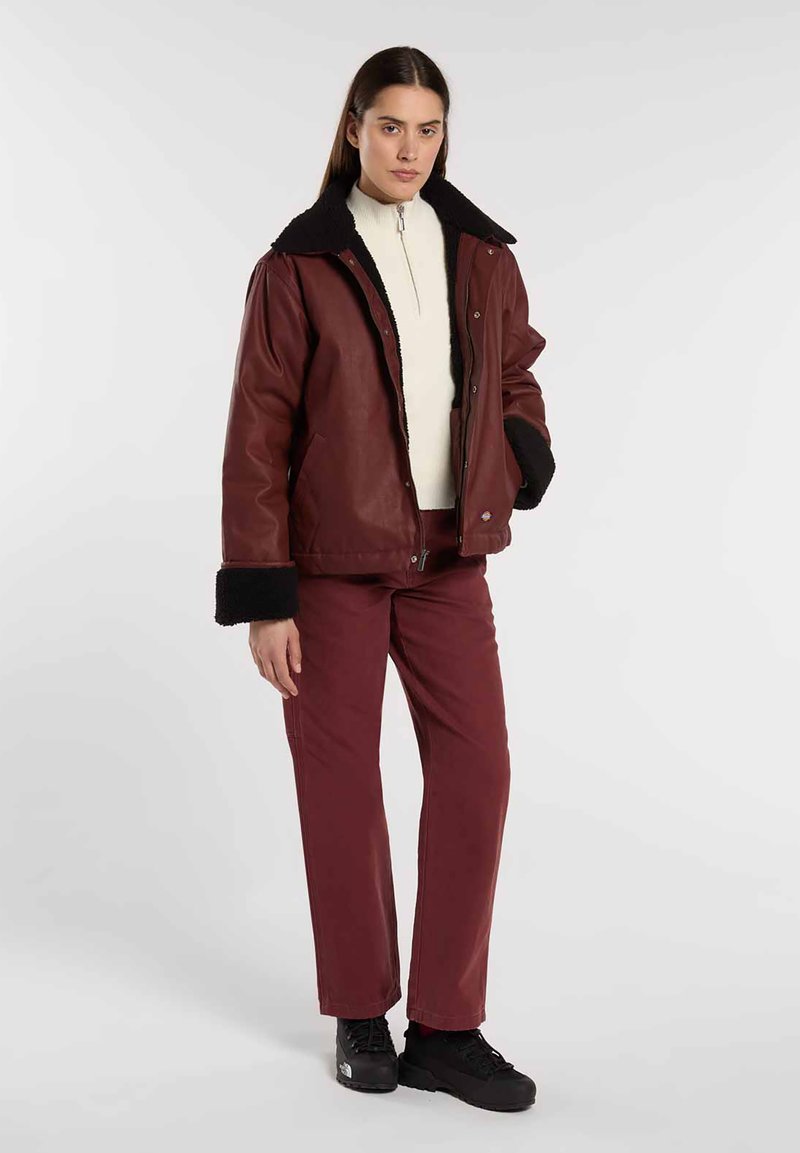 Giacca di pelle rossa con colletto e polsini in shearling nero, indossata sopra un top crema. Abbinata a pantaloni bordeaux e sneakers nere.