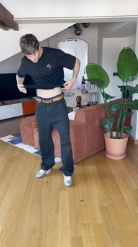 Črna majica in temno jeans hlače z rjavim pasom. Majica ima majhen dizajn na prsih. Prostor je opremljen z lesenim tlakom in zelenimi rastlinami.
