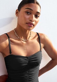Bañador de una sola pieza negro con tirantes finos ajustables, textura fruncida y collar de cadena en tono dorado. La modelo posa frente a un fondo blanco.