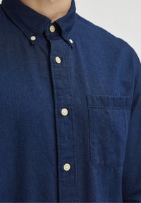 Tmavě modrá košile na knoflíky z denimu, s klasickým límečkem, bílými knoflíky a náprsní kapsou. Hladká textura.