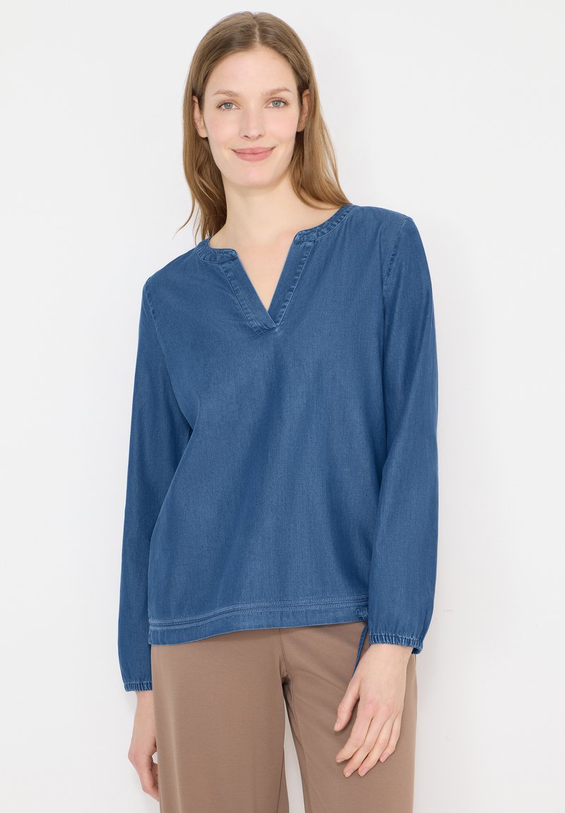 Femme aux cheveux lisses châtain clair, portant un chemisier bleu à manches longues et col en V, ainsi qu'un pantalon beige, debout devant un fond blanc uni.