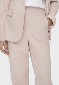 Traje ajustado de color rosa claro que presenta un blazer estructurado y pantalones de pierna ancha con bolsillos laterales y sutiles detalles de costura vertical.