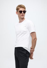 T-shirt bianca a maniche corte con scollo rotondo, abbinata a pantaloni grigio scuro. Il modello indossa occhiali da sole scuri, creando un look casual.