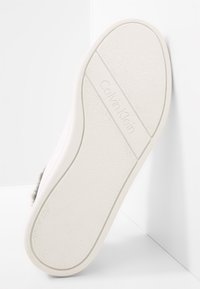 Bílá gumová podrážka s texturovaným povrchem a logem Calvin Klein. Lehce zaoblený tvar s hladkým okrajem a minimalistickým designem.