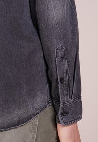 Camisa de mezclilla gris oscuro con puños abotonados y un acabado ligeramente desgastado, con una textura suave y un diseño de cuello clásico.