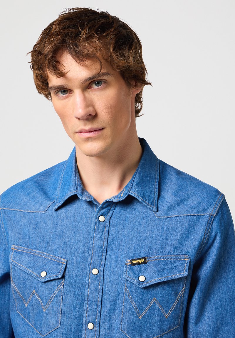 Camisa de mezclilla azul con botones a presión, cuello puntiagudo y dos bolsillos en el pecho con costura en zigzag. Etiqueta del logo en el bolsillo izquierdo.