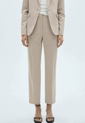 Pantalones beige a medida y blazer a juego con top blanco y sandalias negras de tacón alto y punta abierta, posando contra un fondo liso.