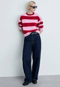 Pull surdimensionné rayé rose et rouge associé à un jean bleu foncé à jambes larges et des chaussures noires. Le pull a un col rond et des manches longues.