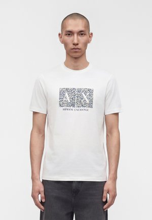 Jeune homme portant un T-shirt blanc à manches courtes avec le motif bleu marine "A X" et le texte "Armani Exchange" sur la poitrine, associé à un pantalon foncé.