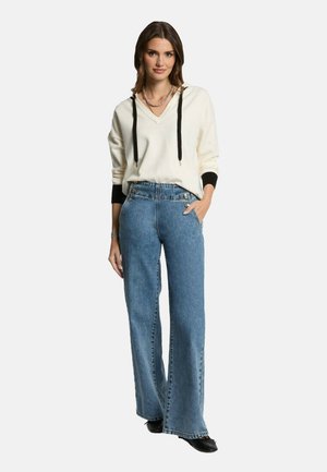 Felpa con scollo a V crema con dettagli neri e cordoncino. Jeans in denim blu chiaro a gamba larga con bottoni frontali. Calzature nere.