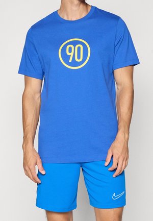 Mann trägt ein blaues T-Shirt mit einem gelben "90" Kreis-Logo und leuchtend blaue Nike-Shorts mit weißem Swoosh-Logo.