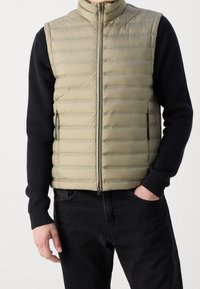 Gilet imbottito beige con cuciture orizzontali, collo alto e due tasche laterali con zip, indossato sopra una maglia nera a maniche lunghe con texture e jeans scuri.
