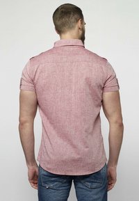 Homme portant une chemise rose à manches courtes et un jean bleu vu de dos sur un fond clair uni.
