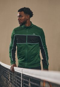 Groene en zwarte sportjas met een rits en zijstrepen, voorzien van een Lacoste-logo. De stof lijkt glad en aansluitend.