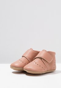 Zapatos de cuero rosa para niños pequeños con tira de velcro, puntera redonda, detalles perforados y suela de goma beige; diseño suave y flexible.