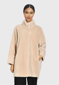 Abrigo de piel sintética beige con cuello alto, corte oversized y bolsillos laterales. Textura suave, botones mínimos y mangas largas.