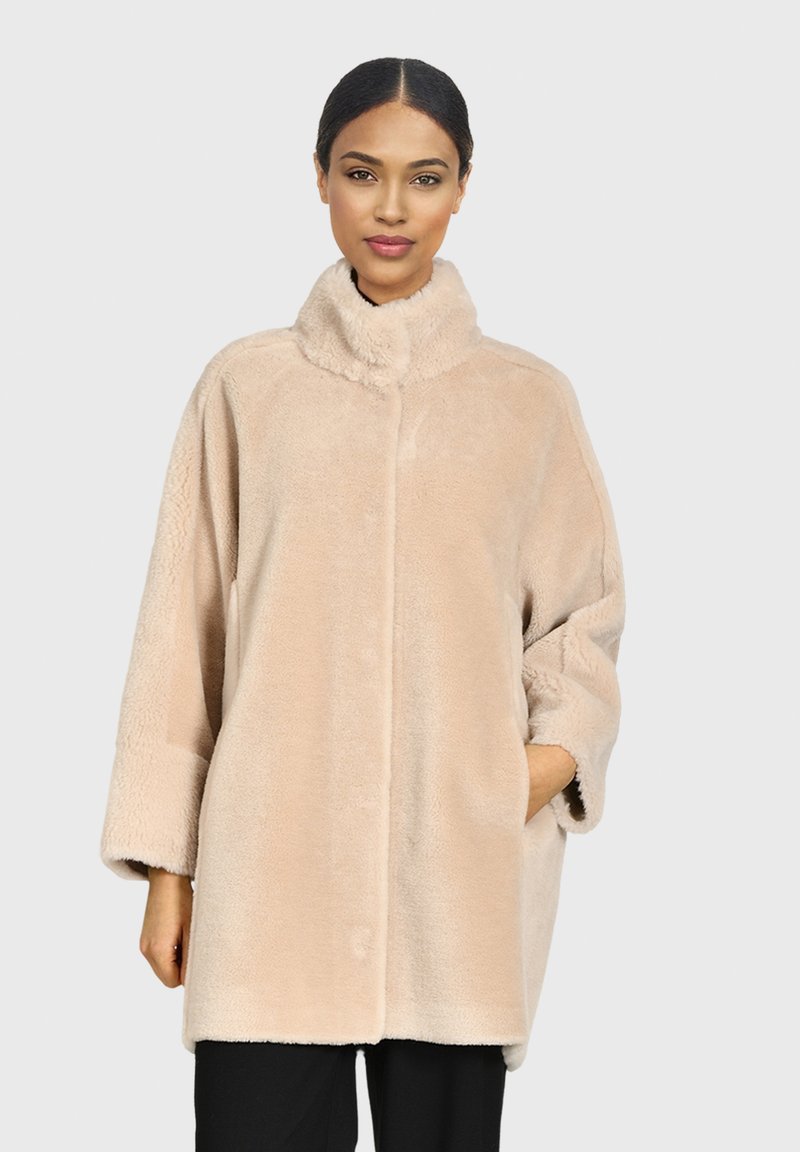 Abrigo de piel sintética beige con cuello alto, corte oversized y bolsillos laterales. Textura suave, botones mínimos y mangas largas.