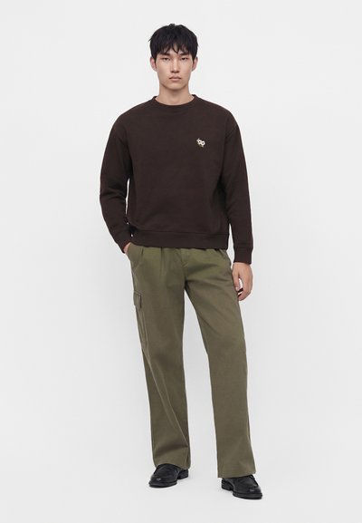 Jeune homme debout portant un sweat-shirt marron foncé avec un petit logo et un pantalon cargo vert olive avec des chaussures noires sur fond blanc.