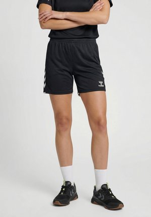 Person står med armene krydset, iført sorte atletikshorts med hvide logoer, sorte sportssko, hvide sokker og en sort top mod en ensfarvet baggrund.