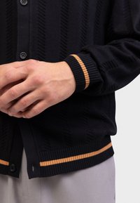 Χέρια που ενώνουν πάνω από ένα μαύρο ζακέτα με κάθετα σχέδια και καφέ ρίγες στα μανίκια και στο τελείωμα, συνδυασμένο με ανοιχτό γκρι παντελόνι.