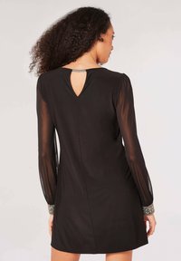 Schwarzes Kleid mit langen, transparenten Ärmeln, Schlitzdetail am Rücken und verzierten Manschetten. Glatter Stoff mit taillierter Silhouette, knielang.